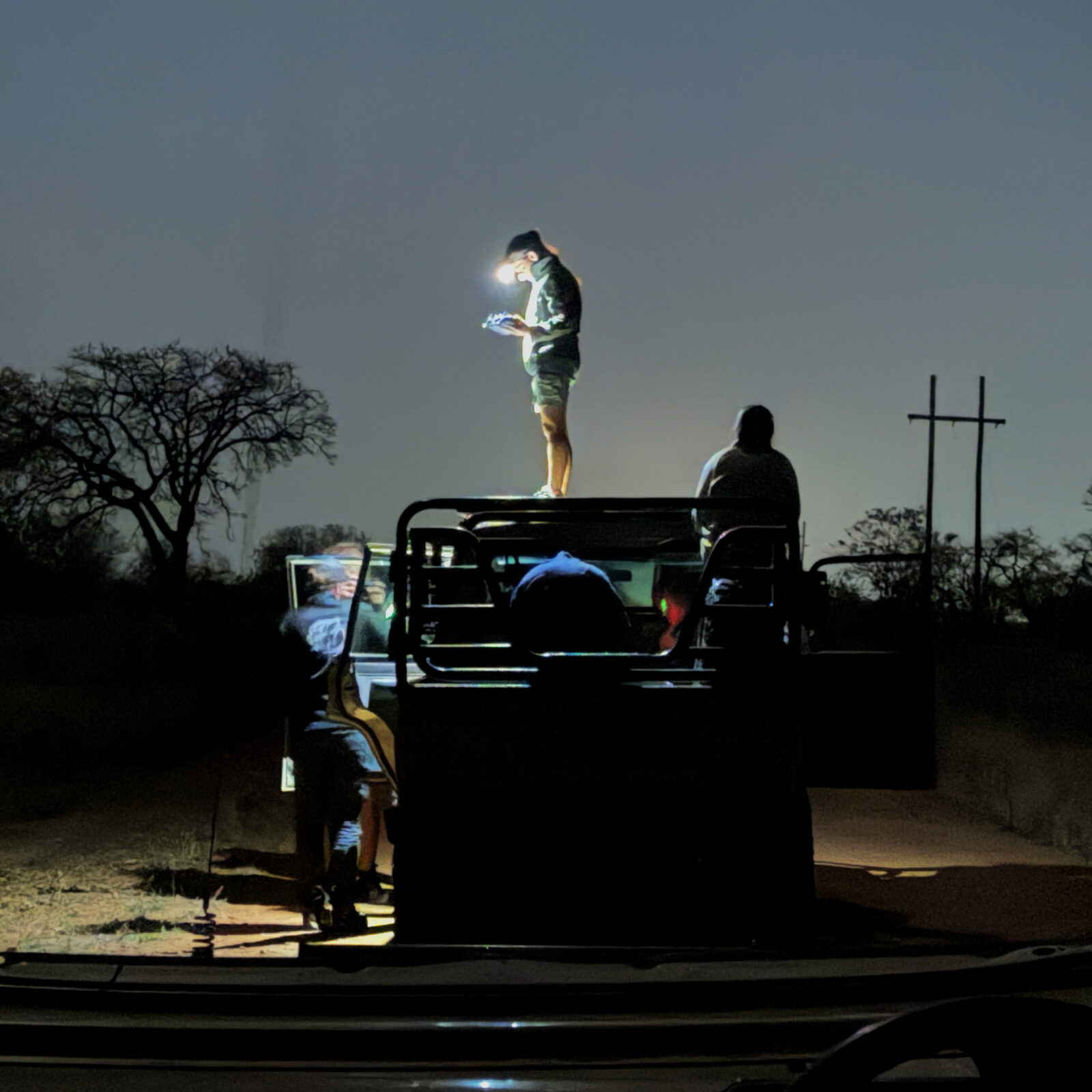 A team member standing on the roof of a vehicle at night, lit by a headlamp and looking at a drone controller
