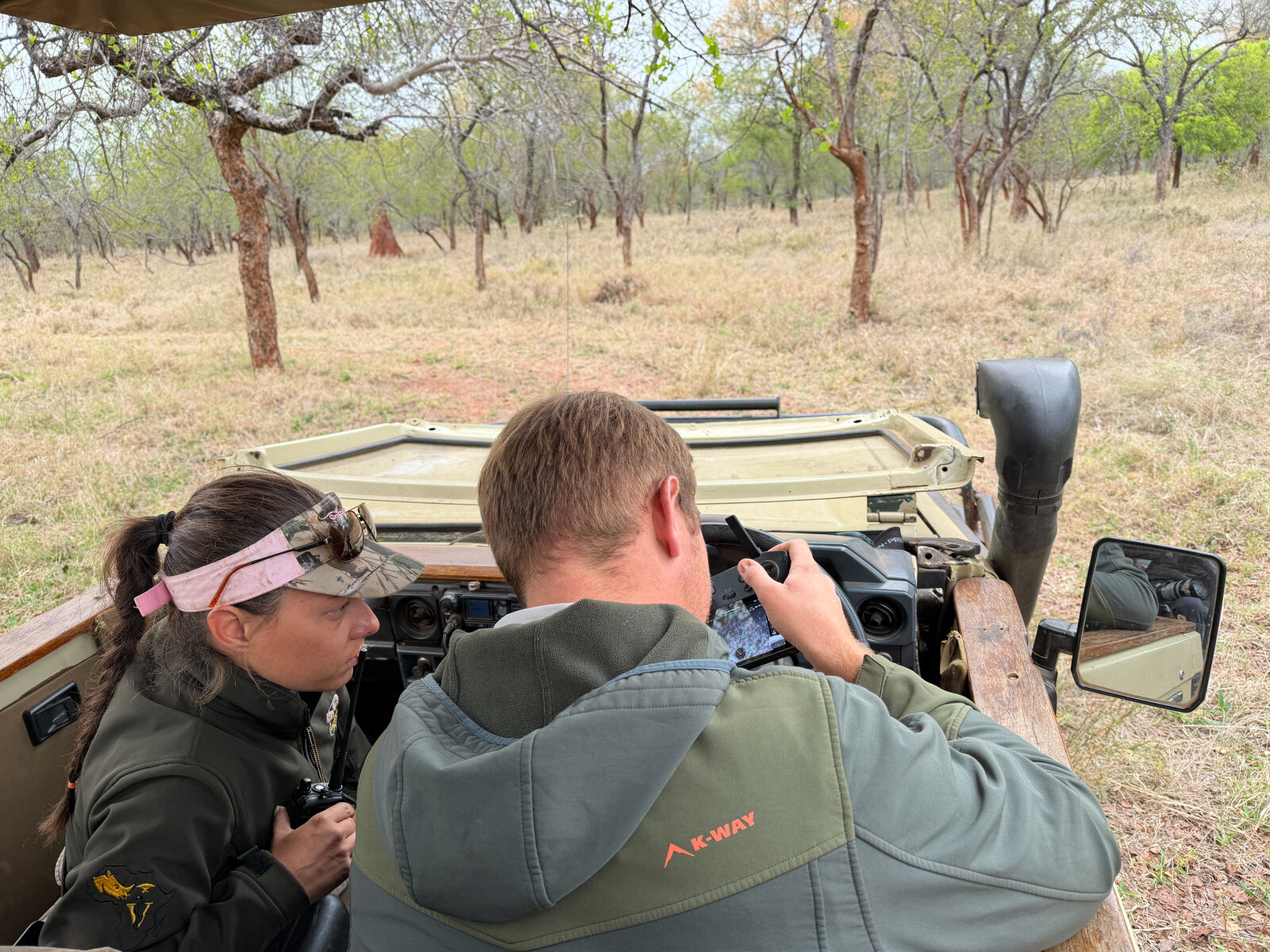 Two team members in an open vehicle looking at a drone controller screen, radio in hand, in dry woodland